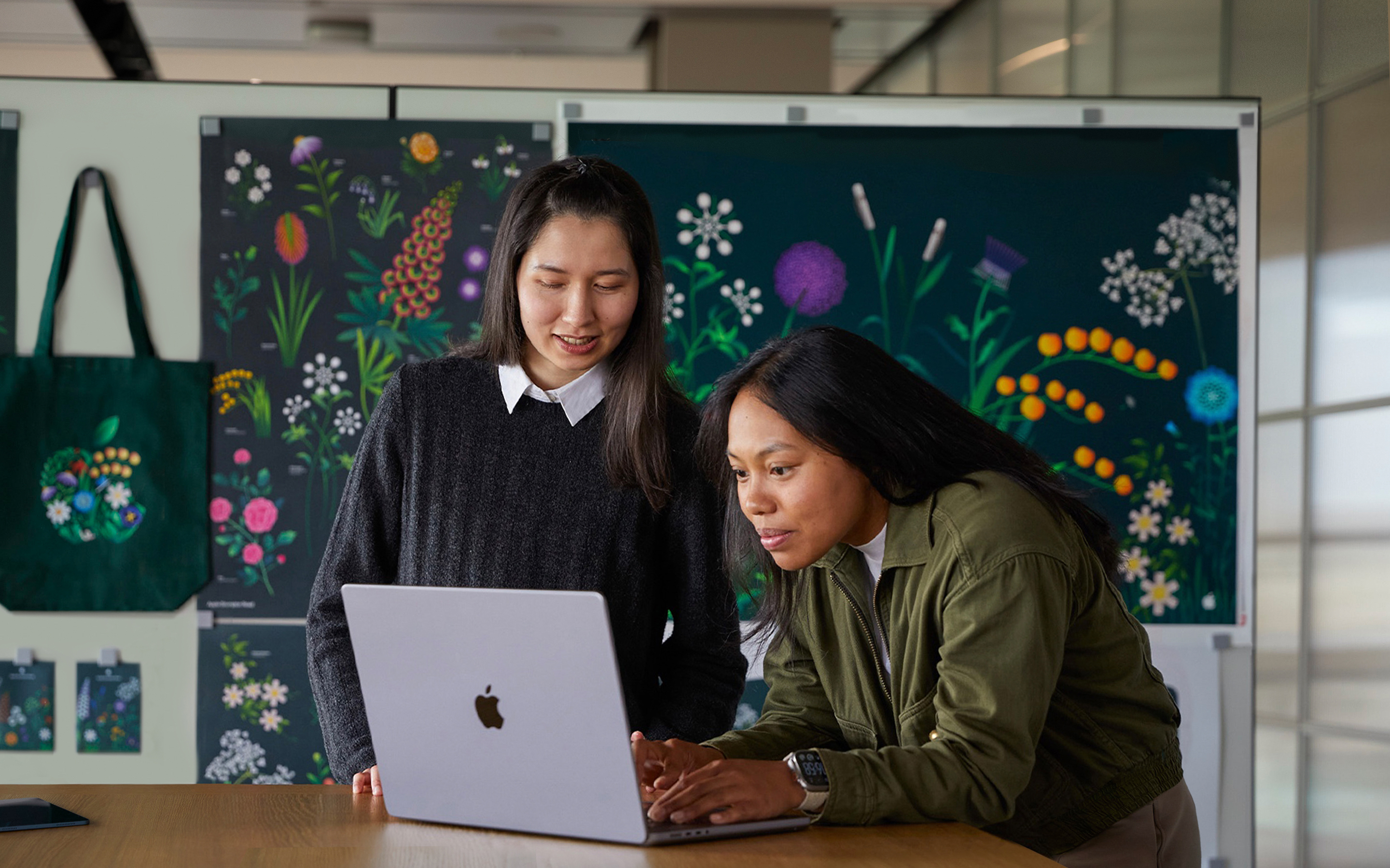 Two Apple Marketing team members working together on a MacBook in front of posters designed by their team.