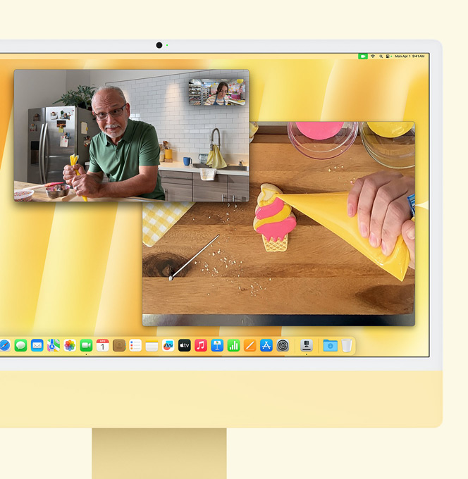 iMac en amarillo. Demostración de Vista Cenital. Se muestra a una panadera haciendo una videollamada con otra persona. Una pantalla muestra a la panadera y el rostro del hombre. La otra muestra las manos de la panadera glaseando una galleta.