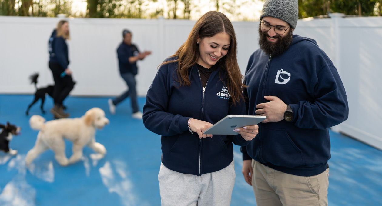 会社のロゴが入ったパーカーを着た2人の社員が、犬の遊び場で一緒にiPadを見ている。背景には犬とスタッフが見える