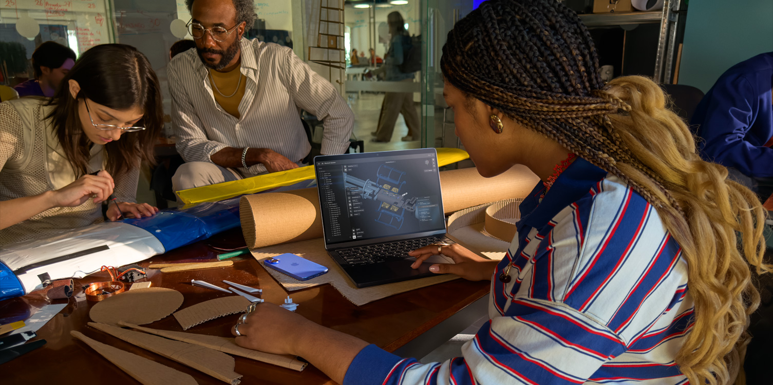 Un grupo de estudiantes sentados en una mesa trabajan en un proyecto técnico y una tiene una MacBook abierta en la que se ve la app Shapr 3D.