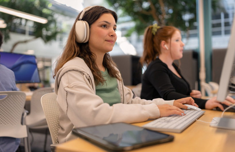 Uma estudante usando os AirPods Max está sentada a uma mesa com um computador e digita em um teclado, com um tablet ao lado. Outra estudante trabalha ao lado dela