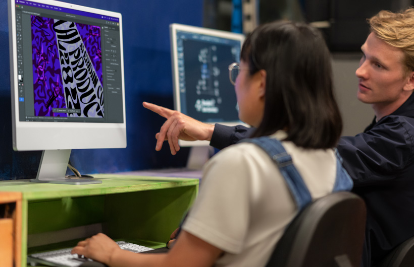 Dois estudantes sentados em frente a telas de iMac que mostram modelos coloridos. Um dos estudantes aponta para a tela