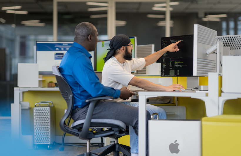 Dois estudantes sentados na frente de várias telas de iMac. Um dos estudantes aponta para uma tela