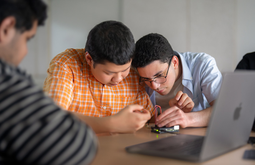 Dois estudantes observam atentamente uma placa de circuito ao lado de um MacBook, enquanto um terceiro olha na direção deles