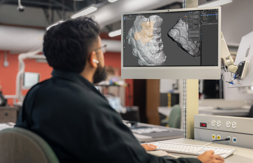 Um estudante sentado à mesa revisa dois modelos de cabeça escaneados em 3D exibidos lado a lado em telas do iMac