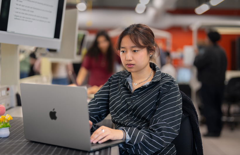 Uma estudante sentada à mesa em uma sala de aula moderna usa um MacBook