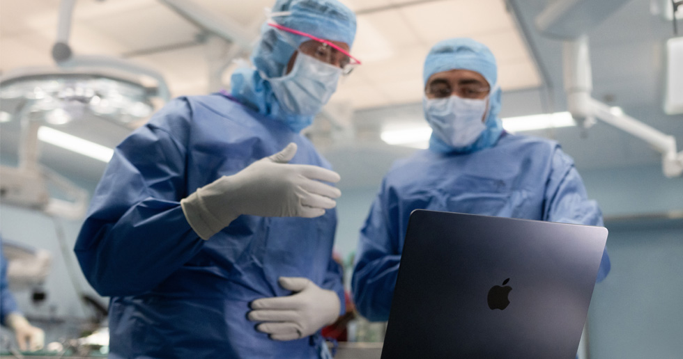To kirurger i et operasjonsrom ser på en MacBook, og den ene gestikulerer mens den andre ser på