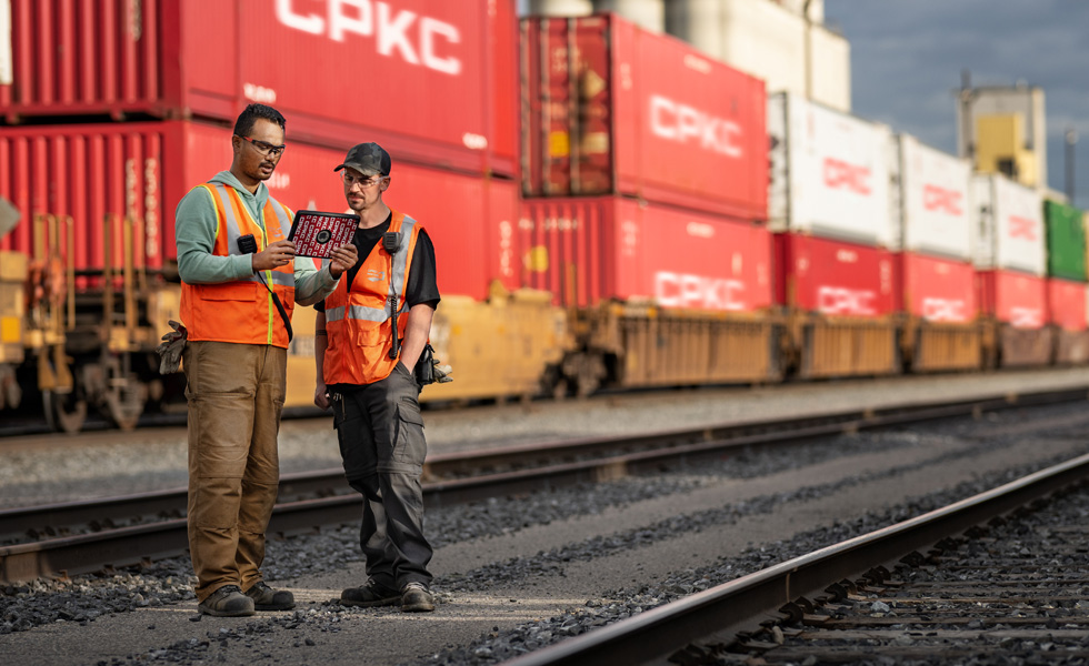 Twee spoorwegmedewerkers gebruiken een iPad terwijl ze op een rangeerterrein naast een stilstaande trein met zeecontainers staan