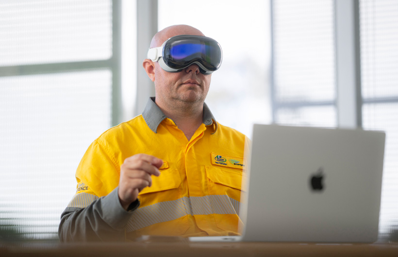 Funcionário com um Apple Vision Pro a fazer o gesto de juntar o polegar e o indicador, com um MacBook Pro prateado aberto na secretária