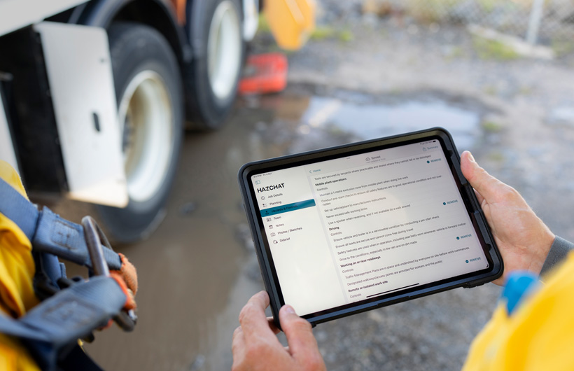 Mãos a segurar um iPad com a app HazChat aberta, que apresenta as opções Detalhes do trabalho, Planeamento, Perigos e controlos, e uma pessoa num estaleiro junto a uma poça de lama