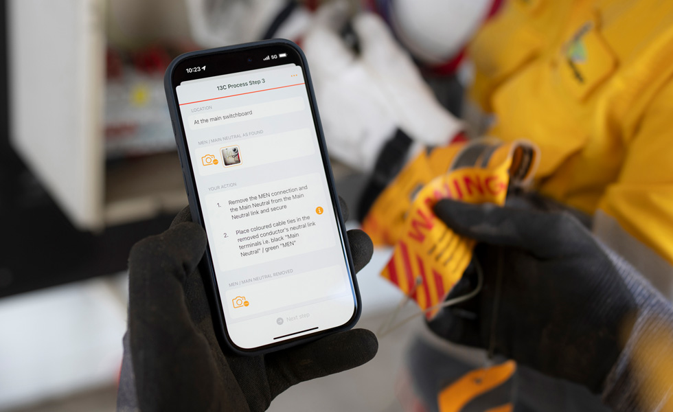 Mãos a segurar um iPhone que apresenta instruções para fazer um teste do quadro elétrico, com o título "13C Process Step 3" no ecrã