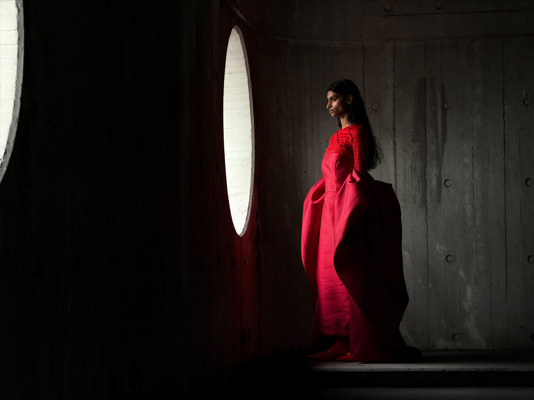 Seorang perempuan memakai gaun merah panjang dalam ruangan kosong sedang difoto. Aplikasi Foto beralih antar jarak fokus mulai dari 200 mm hingga 13 mm, menunjukkan kemampuan sistem kamera Pro
