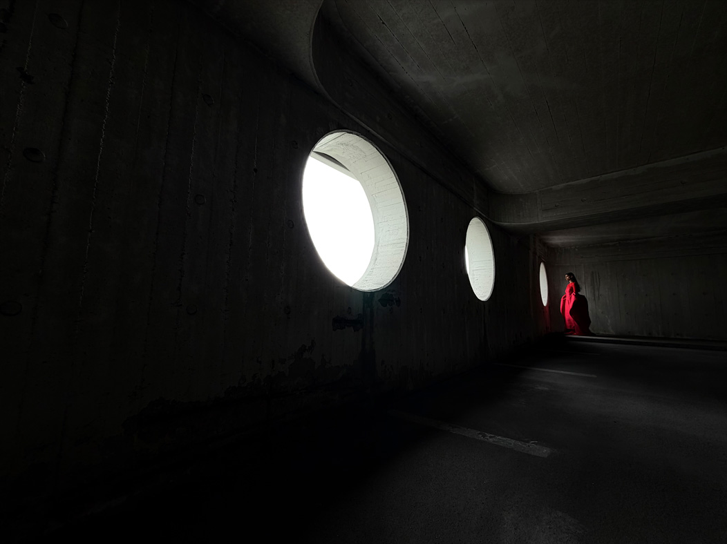 Seorang perempuan memakai gaun merah panjang dalam ruangan kosong sedang difoto. Aplikasi Foto beralih antar jarak fokus mulai dari 200 mm hingga 13 mm, menunjukkan kemampuan sistem kamera Pro