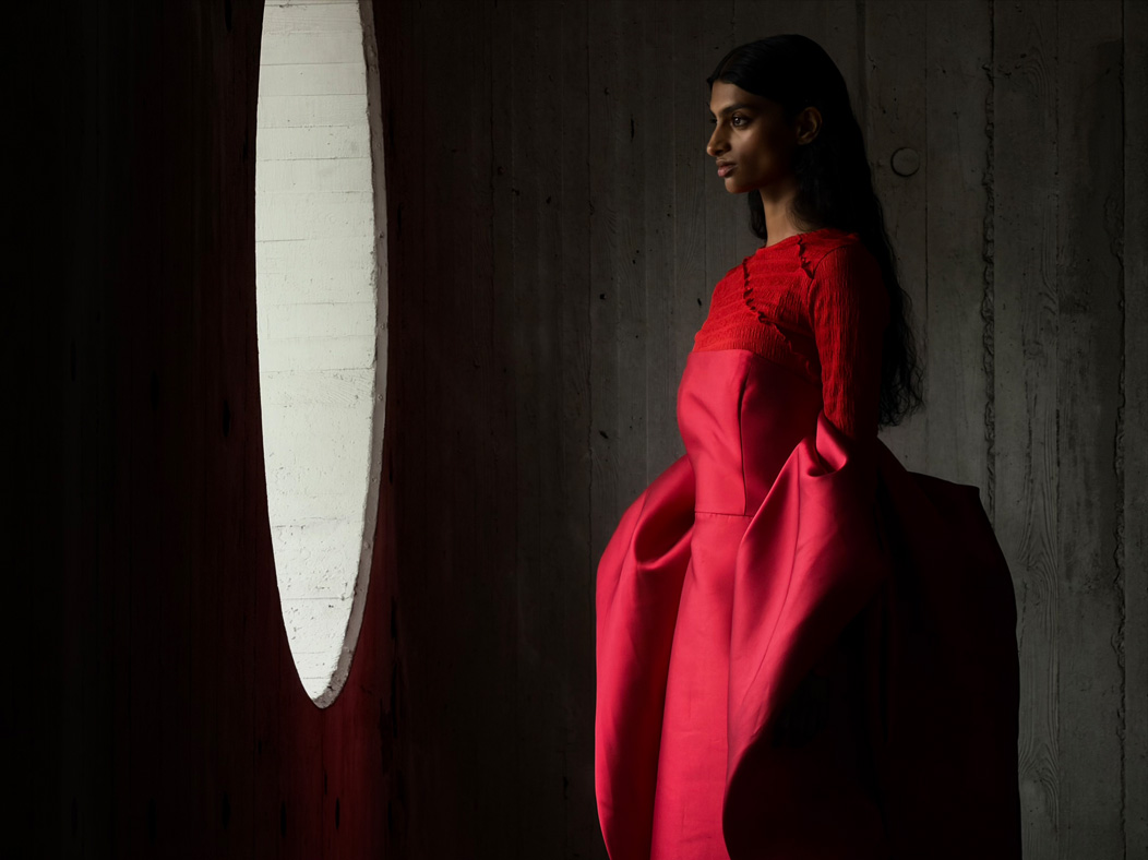 Seorang perempuan memakai gaun merah panjang dalam ruangan kosong sedang difoto. Aplikasi Foto beralih antar jarak fokus mulai dari 200 mm hingga 13 mm, menunjukkan kemampuan sistem kamera Pro